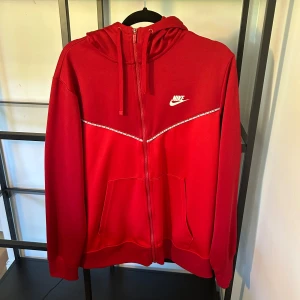 Röd Nike hoodie med dragkedja - Nike hoodie i rött med vit logga på bröstet och snygg detalj över bröstet. Tröjan har huva med snörning, hel dragkedja och två stora fickor framtill. Perfekt för en sportig och avslappnad look.