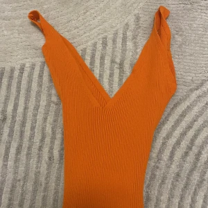 Ribbad orange klänning med knytband - Säljer en ribbad orange klänning med v-ringning och smala axelband. Klänningen har ett matchande knytband i midjan som ger en snygg siluett. Modellen är figurnära och materialet är stretchigt och bekvämt. Perfekt för dig som vill sticka ut med färg!