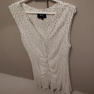 Vit prickig klänning från Object - Snygg vit klänning från Object med svarta prickar, v-ringad hals och knappar framtill. Klänningen är ärmlös och har en rynkad detalj längs magen som ger en cool form. Materialet känns mjukt och lätt, perfekt för varmare dagar.