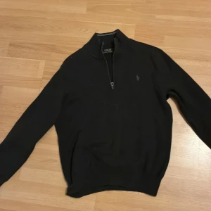 Svart stickad tröja Polo Ralph Lauren - Svart stickad tröja från Polo Ralph Lauren med halv dragkedja och klassisk broderad logga på bröstet. Tröjan har lång ärm och ribbade muddar, perfekt för lager-på-lager under kyliga dagar.