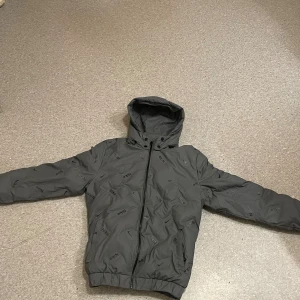 Grå pufferjacka från Nike med huva - Snygg grå pufferjacka från Nike med svart logotypmönster över hela jackan. Jackan har huva, dragkedja framtill och elastiska muddar vid ärmslut och midja. Perfekt för kyliga dagar och ger en sportig vibe. Men vänster ficka är trasig därav priset! 