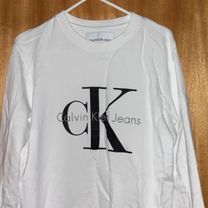 Vit långärmad tröja Calvin Klein Jeans - Vit långärmad tröja från Calvin Klein Jeans med stor svart CK-logga och text framtill. Tröjan har rund halsringning och är gjord i mjuk bomull, perfekt för en clean och stilren look.