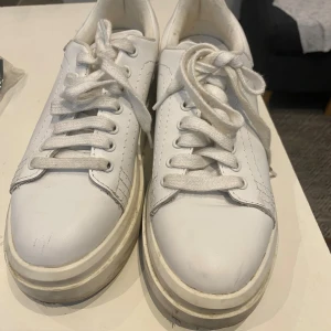 Vita sneakers med platåsula - Säljer ett par helvita sneakers med platåsula och snörning. Skorna har rund tå och är tillverkade i syntetmaterial med tydliga sömmar och klassisk design. Perfekta för dig som gillar en clean och stilren look.