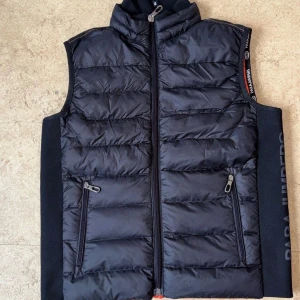  Parajumpers väst  - Snygg svart dunväst från Parajumpers med quiltad design och orange foder. Västen har dragkedja framtill, två fickor med dragkedja och tydlig logga på sidan. Perfekt för lager-på-lager och har en sportig vibe.