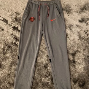 Grå Nike PSG mjukisbyxor  - Grå mjukisbyxor från Nike med Paris Saint-Germain-logga och röd Nike Swoosh på framsidan. Byxorna har snörning i midjan, ribbade muddar och svarta banddetaljer på baksidan av benen. Perfekta för chill eller sport.