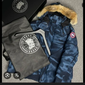 Blå camo dunjacka från Canada Goose - Säljer en sällsynt  blå camo dunjacka från Canada Goose med camo-liknande mönster och ikonisk patch på ärmen. Jackan har pälskantad huva, dragkedja och knappar framtill. Perfekt för kalla vinterdagar och riktigt snygg streetstil.