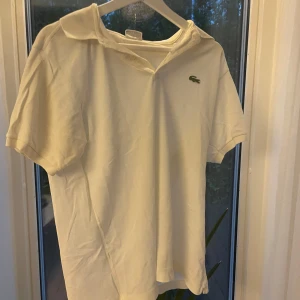 Vit pikétröja från Lacoste - Säljer en klassisk vit pikétröja från Lacoste med den ikoniska gröna krokodilen broderad på bröstet. Tröjan har korta ärmar, krage och är gjord i mjuk bomull som är skön mot huden. Perfekt för dig som gillar stilrena och tidlösa plagg.