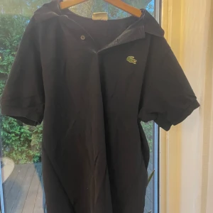 Svart/grå pikétröja från Lacoste - Klassisk grå pikétröja från Lacoste med korta ärmar och den ikoniska gröna krokodilloggan på bröstet. Tröjan har krage och knäppning framtill. Perfekt för en stilren och sportig look.