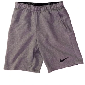 Grå shorts från Nike - Snygga grå shorts från Nike med elastisk midja och två sidofickor. Klassisk svart Nike-logga på ena benet. Perfekta för träning eller chill, tillverkade i mjuk bomullsblandning för maximal komfort.