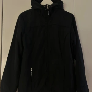 Svart vindjacka med huva - Svart vindjacka med huva och dragkedja framtill. Jackan har två sidofickor med dragkedja och en enkel, stilren design. Perfekt för blåsiga dagar och har en normal passform med långa ärmar. Materialet är troligtvis polyester. Inhandlades för 500 kr - men jag säljer för billigare!✨
