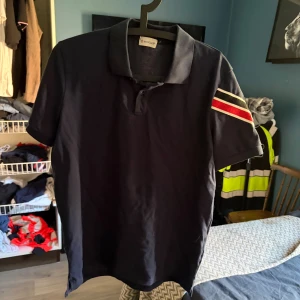 Mörkblå Moncler pikétröja med ränder - Snygg mörkblå pikétröja från Moncler med klassisk krage och korta ärmar. På ena ärmen finns en bred rand i rött, vitt och svart samt Moncler-logga. Tröjan är i mjuk bomull och har en stilren look som passar perfekt till jeans eller shorts.