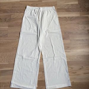Vita linnebyxor med dragsko - Luftiga vita linnebyxor med resår och snörning i midjan. Byxorna har raka ben, sidfickor och en avslappnad passform. Perfekta för varma dagar. Materialet är lätt och har en lite skrynklig struktur. Det finns en liten fläck på vänster ficka, har tyvärr inte testat att tvätta. 