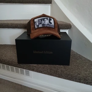 Brun keps från Madoura Limited Edition - Säljer en brun keps från Madoura Limited Edition med vintageinspirerad svartvit patch framtill som visar ett coolt motiv. Kepsen har klassisk form med böjd skärm och mesh på sidorna för extra ventilation. Perfekt accessoar för en unik streetstyle.