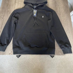 Svart hoodie från Polo Ralph Lauren - Svart hoodie från Polo Ralph Lauren med klassisk huva och snörning. Tröjan har en stor magficka och den ikoniska lilla vita Polo-loggan broderad på bröstet. Tillverkad i mjuk bomull, perfekt för chill dagar. Sitter som en s