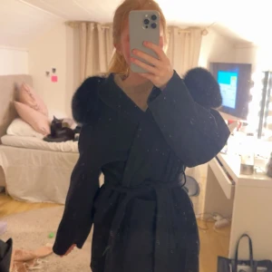 Svart kappa med fuskpälsdetaljer - Säljer en svart kappa med fusk päls på luvan och knytskärp i midjan. Jackan har en klassisk, rak passform och är perfekt för dig som vill sticka ut lite extra. Enkel att styla och supermysig under kyliga dagar.