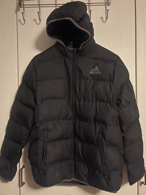 Svart pufferjacka från Adidas - Svart pufferjacka från Adidas med huva och dragkedja framtill. Jackan har två fickor med dragkedja och Adidas-logga på bröstet. Perfekt för kalla dagar och har en sportig vibe. Materialet är syntet och jackan är vadderad för extra värme. Passar tonåring eller vuxen ca 160-165 cm lång. Enligt tillverkarens storlek: 13-14 år. Eller 164 cl.