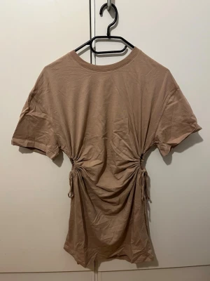 Beige kort klänning från Bershka - Säljer en beige kort klänning från Bershka med snörning i midjan som ger en snygg draperad effekt. Klänningen har rund hals och korta ärmar, tillverkad i mjuk bomull. Använd ca. 2-3 gånger så är i princip i nyskick