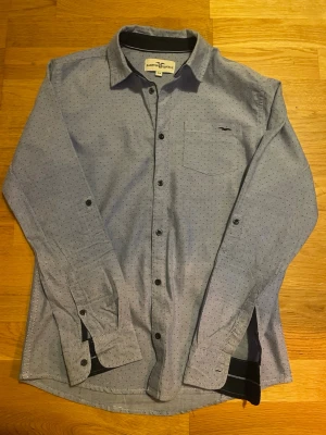 Blå prickig skjorta Hampton Republic - Snygg blå skjorta från Hampton Republic med små svarta prickar och klassisk krage. Skjortan har bröstficka med broderad logga, långa ärmar och knappar framtill. Perfekt till jeans eller chinos för en clean och stilren look. I barnstorlek 164