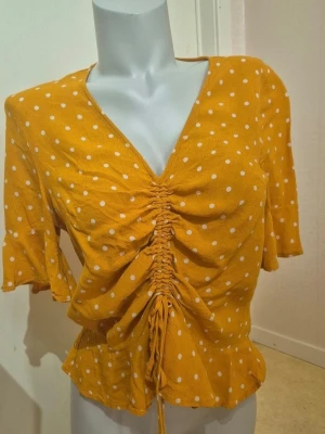 Gul prickig blus från H&M - Säljer en gul blus från H&M med vita prickar, v-ringad och snörning framtill som ger en snygg rynkad effekt. Blusen har korta ärmar med volangkant och är croppad i modellen. Perfekt för dig som gillar färg och mönster!💛🤍🛍