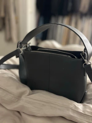 Svart axelväska i skinn med dragkedja - Snygg svart axelväska i skinn med stilren design och silverfärgad dragkedja. Väskan har ett brett axelband och flera fack inuti för smart organisering. Perfekt storlek för att få plats med allt viktigt och passar till många olika stilar.