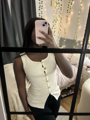 Vit ärmlös topp med guldknappar - Snygg vit ärmlös topp med raka axelband och dekorativa guldknappar längs hela framsidan. Toppen har en figurnära passform och är perfekt att styla med både jeans och kjol. Materialet känns mjukt och stretchigt, vilket gör den bekväm att bära hela dagen.