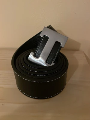 Svart skinnbälte med metallspänne - Snyggt svart bälte i skinn med ett stort, silvrigt metallspänne format som ett H. Bältet har vita kontrastsömmar längs kanterna som ger en cool detalj. Perfekt för att lyfta din outfit med en stilren accessoar.