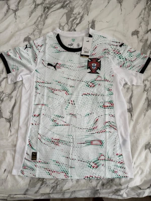 Vit Portugal fotbollströja Puma - Säljer en vit Portugal fotbollströja från Puma med coolt prickigt mönster i rött och grönt. Tröjan har svarta detaljer vid krage och ärmslut samt Portugal-emblem på bröstet. Perfekt för dig som älskar fotboll och vill ha en snygg supportertröja.