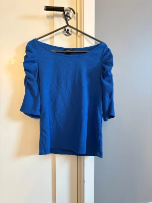 Blå topp med rynkade ärmar från Lindex - Säljer en blå topp från Lindex i storlek XS. Toppen har rynkade korta ärmar. Modellen är rak med rundad halsringning. 