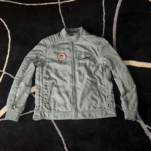 Grön bomberjacka från Hampton Republic - Snygg grön bomberjacka från Hampton Republic med coolt emblem på bröstet och mönstrat innerfoder.  Jackan har dragkedja framtill, fickor med dragkedja och en extra ficka med knapp på bröstet.  Perfekt för dig som gillar streetstyle och vill ha en jacka med attityd. Jackan har tyvärr några fläckar vid höger axel (se bilderna). Storlek L.