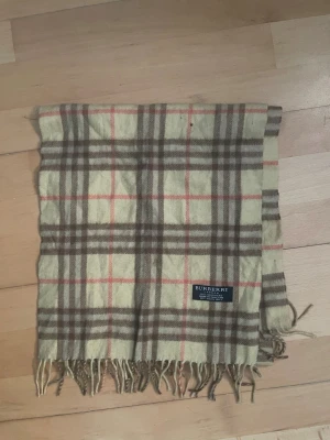 Rutig beige Burberry halsduk i kashmir - Säljer en klassisk rutig halsduk från Burberry i 100% kashmir. Halsduken har beige botten med bruna och röda rutor, samt fransar längst ner. Märket sitter tydligt på framsidan. Perfekt för dig som gillar tidlös design och mjuka material. Längd 135 bredd 30