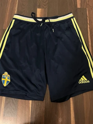 Shorts  - Bilden visar ett par mörkblå shorts från Adidas med gula detaljer och SVFF-logga! 