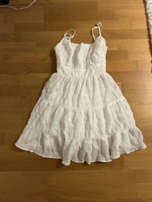 Vit omlottklänning med volanger - Säljer en söt vit kortklänning med smock upptill och tunna axelband som kan knytas. Klänningen har volanger och ett omlottliknande fall i kjolen, vilket ger en luftig och somrig känsla. Materialet är mjukt och lätt, perfekt för varma dagar. Köptes second hand och säljer vidare den nu då den ej används längre