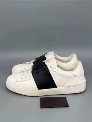 Valentino Garavani Open sneakers vit/svart - Säljer ett par Valentino Garavani Open sneakers i vitt skinn med bred svart läderpanel över snörningen. Skorna har rund tå, platt sula och dekorativa nitar på hälen. Snygg kontrast mellan vit och svart, perfekt för dig som gillar stilrena designer.