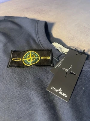 Mörkblå sweatshirt från Stone Island - Snygg mörkblå sweatshirt från Stone Island med klassisk logotyp-patch på vänster ärm. Tröjan har rund hals, ribbade muddar och är tillverkad i mjuk bomull. Perfekt för dig som gillar streetwear och vill ha en clean look med tydlig branding.
