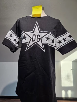 Svart DG t-shirt med stjärnor - Cool svart t-shirt från DG med stort vitt stjärntryck och DG-logga på bröstet. T-shirten har korta ärmar och ett brett band med svarta stjärnor över bröstet och ärmarna. Perfekt för dig som gillar streetwear och grafiska tryck.
