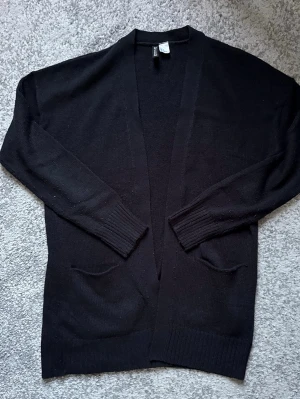 Svart kofta från H&M - Säljer en svart, långärmad kofta från H&M med två praktiska fickor framtill. Koftan är stickad och har ribbade muddar vid ärmslut och nederkant. Perfekt att slänga över en t-shirt eller linne för en chill look.