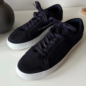 Marinblåa sneakers från Les Deux i mocka - Snygga marinblåa sneakers från Les Deux med ovandel i mjuk mocka och vita platta sulor. Skorna har snörning och broderade detaljer i blått, vitt och rött på hälen. Perfekta för dig som gillar stilrena och klassiska sneakers med en twist.