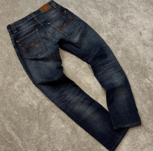 Mörkblå Grim Tim jeans  från Nudie Jeans - Snygga mörkblå Grim Tim jeans från Nudie Jeans med klassisk femficksdesign och orangea sömmar. Jeansen är tillverkade i slitstarkt denim med coola slitningar på framsidan och bakfickor med Nudie-signatur. Perfekt för dig som gillar en tidlös look.