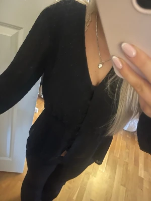 Svart blus med v-ringning - Säljer en svart blus med v-ringning och långa ärmar. Blusen har en lätt volang vid midjan och knappar framtill. Materialet är tunt och ger en luftig känsla, perfekt för dig som gillar stilrena och enkla plagg.