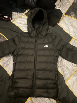 Svart pufferjacka från Adidas - Svart pufferjacka från Adidas med huva och dragkedja framtill. Jackan har ett stilrent Adidas-logga på bröstet och är quiltad för extra värme. Perfekt för kalla dagar och har en modern, loose passform som är både bekväm och snygg.