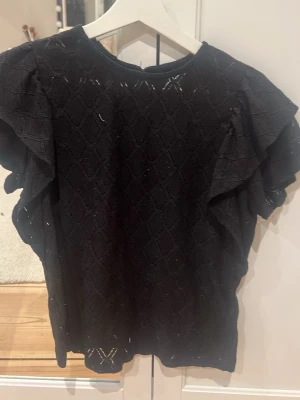 Svart blus med volangärmar - Säljer en svart blus med snyggt strukturmönster och volangdetaljer på ärmarna. Blusen har rund halsringning och knapp i nacken. Materialet känns som bomull och har ett diskret rutmönster. Perfekt för dig som gillar stilrena och trendiga toppar.