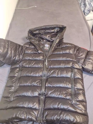 Svart dunjacka från Canada Goose - Säljer en svart, glansig dunjacka från Canada Goose med huva och två stora dragkedjefickor framtill. Jackan har det klassiska röda Canada Goose-märket på ärmen och reflekterande detaljer på huvan. Perfekt för kalla dagar och har en riktigt schysst look.