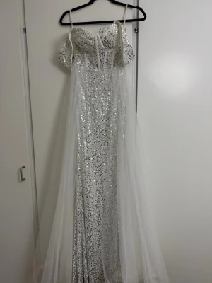 Glittrig långklänning med paljetter - Säljer en magisk långklänning i vit och silver med glittriga paljetter över hela klänningen. Klänningen har tunna axelband, offshoulder-detaljer och ett lager av tunn tyll som ger extra volym. Perfekt för dig som vill sticka ut med en glamorös look.