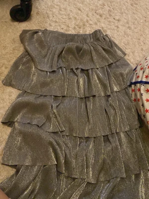 Glittrig volangkjol från NA-KD - Säljer en glittrig silverfärgad kjol från NA-KD i storlek S. Kjolen har flera lager med volanger och ett elastiskt midjeband. Materialet är syntet och har en skimrande finish som verkligen sticker ut. Perfekt för dig som gillar att synas och vill ha något extra till garderoben.