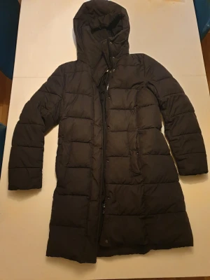 Svart lång pufferjacka från H&M - Säljer en svart lång pufferjacka från H&M i storlek S. Jackan har huva, dragkedja och knappar framtill samt quiltad design. Perfekt för kalla dagar och riktigt skön att bära. Passar dig som vill ha en varm och stilren vinterjacka.
