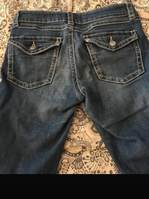 Blå bootcut jeans  - Snygga blå jeans med bootcut passform och kontrasterande vita sömmar. Jeansen har dekorativa bakfickor med knappdetaljer och klassisk femficksdesign. Materialet är slitstarkt jeans och midjan är normal.