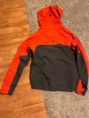 Orange och grå Marmot Gore-Tex vindjacka - Säljer en Marmot vindjacka i Gore-Tex med schysst orange och grå färgblock. Jackan har huva, dragkedjor på fickor och ärmar, samt justerbara muddar. Perfekt för dig som gillar att vara ute oavsett väder. Marmot-logga på bröstet och Gore-Tex-tryck på ärmen.