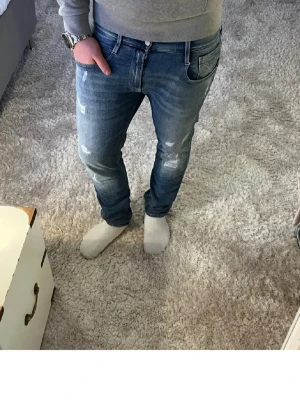 Replay blå jeans med slitningar - Säljer ett par snygga Replay jeans i blå tvätt med coola slitningar och klassisk femficksdesign. Jeansen har normal passform och raka ben, med en tydlig Replay-logga på bakfickan. Perfekt för dig som gillar en avslappnad och trendig stil. Storlek 31/32