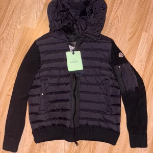 Svart Moncler Cardigan med huva - Säljer en svart Cardigan från Moncler med stickade ärmar och vadderad framsida. Jackan har huva, dragkedja framtill och fickor med knapp. Moncler-logga på ena ärmen och snygga detaljer i både stickat och syntetmaterial.