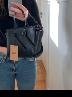 Svart handväska  - Snygg svart handväska med stilren design och två handtag. Väskan har dekorativa detaljer framtill och är tillverkad i skinnimitation. Perfekt storlek för det viktigaste och passar till jeans eller kappa. En modern accessoar som lyfter din look.